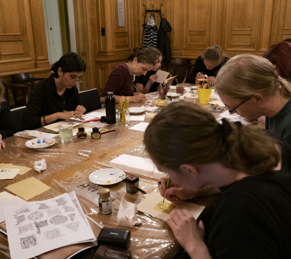 Mehrere Personen sitzen an einem großen Tisch und malen auf Pergament.