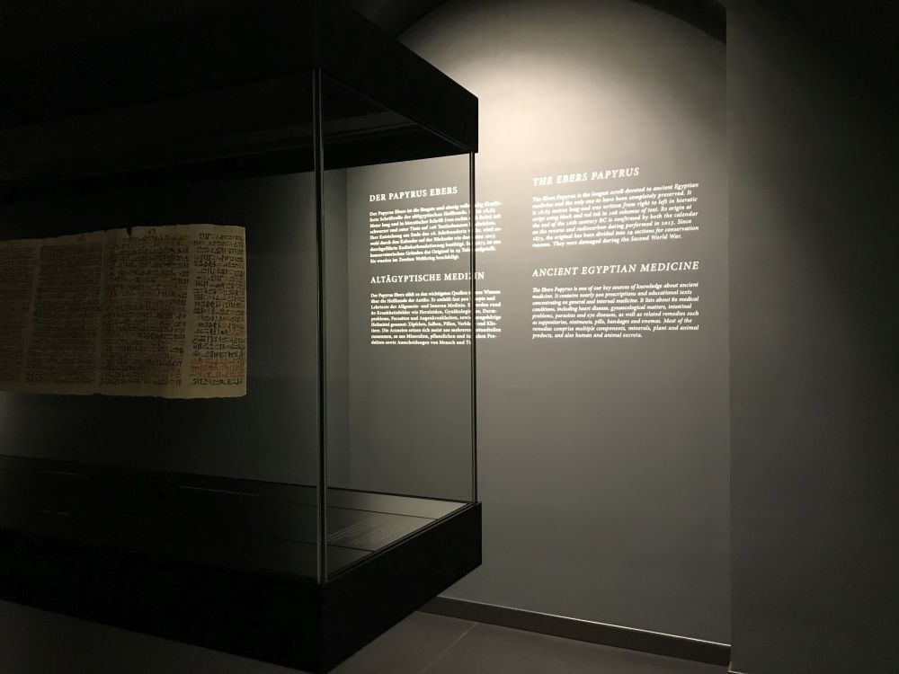 Ein Raum mit schwarzen Wänden und einer Glasvitrine. In der Vtrine ist ein Papyrus ausgestelt, an den Wänden sind weiße Beschreibungstexte auf deutsch und englisch zu sehen.