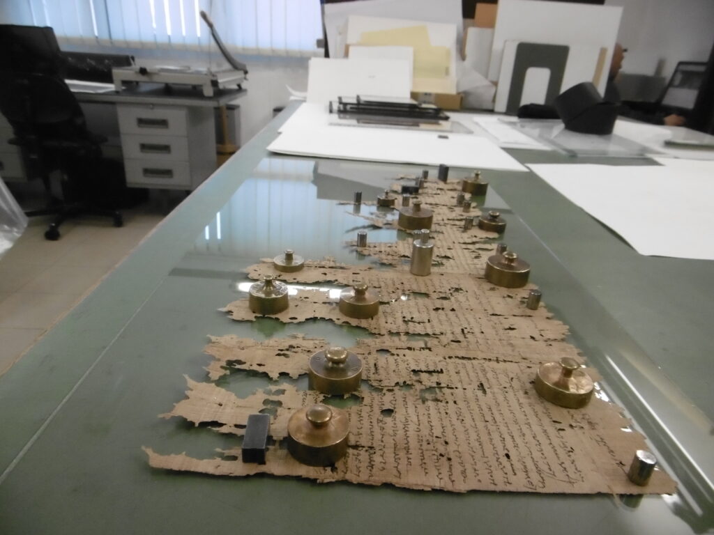 Papyrusfragmente liegen auf einem Tisch. Sie sind mit kleinen Gewichten beschwert.